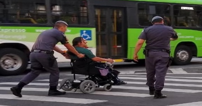 Policiais ajudam cadeirante a atravessar a rua.